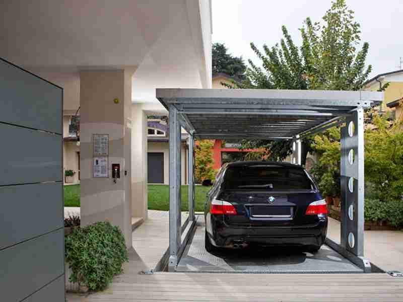 Black car parked under grey metal structure with roof, garden and house in the background